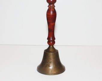 May include: A vintage brass bell with a dark brown wooden handle. The bell has an engraved floral design on its surface. The handle is a rich, polished wood, and the bell is a warm, antique brass color.