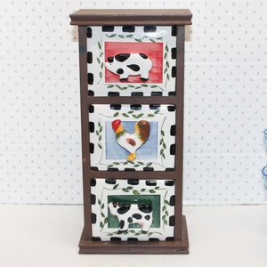 May include: A wooden three-drawer storage box with a black and white checkered pattern. Each drawer features a different colorful ceramic tile with a farm animal design: a pig, a rooster, and a cow.