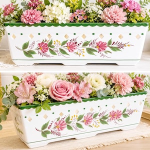 May include: A rectangular white ceramic planter filled with an assortment of pink, white, and yellow flowers. The planter has a green scalloped rim and is decorated with pink floral designs and green leaves. The planter is approximately 12 inches long.