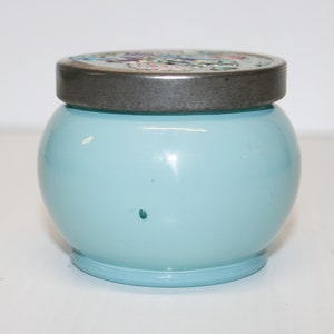 May include: A round, light blue jar with a decorative lid. The lid is silver-toned with a floral design. The jar has a smooth, glossy finish and sits on a small, matching base. The jar is approximately 4 inches tall.