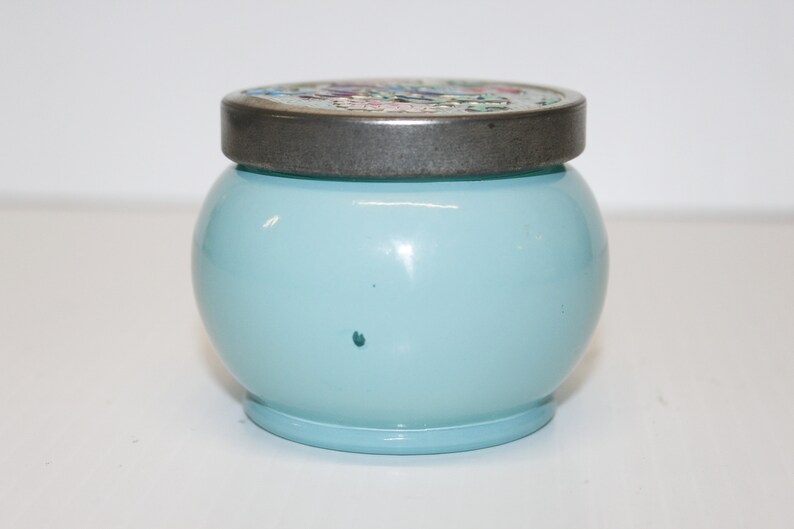 May include: A round, light blue jar with a decorative lid. The lid is silver-toned with a floral design. The jar has a smooth, glossy finish and sits on a small, matching base. The jar is approximately 4 inches tall.