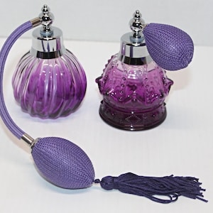 May include: Two vintage purple glass perfume bottles with silver accents. One has a ribbed design, the other a decorative pattern. Both have a bulb atomizer, a matching purple cord and a tassel.