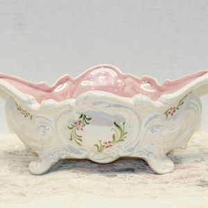 May include: A white ceramic bowl with a pink interior and floral designs. The bowl has a scalloped edge and three feet.