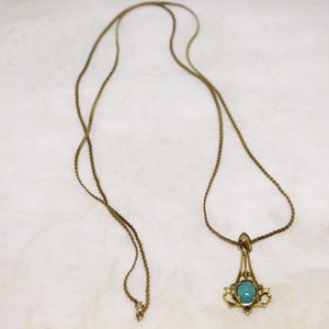 May include: A vintage gold-tone necklace with a pendant featuring a light blue oval stone. The pendant has an ornate, filigree design. The necklace chain is a delicate, woven style.