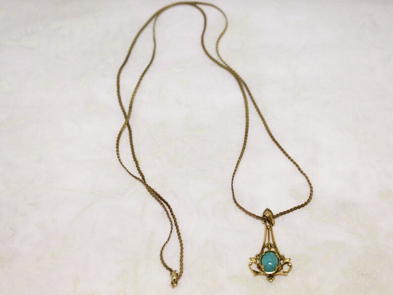 May include: A vintage gold-tone necklace with a pendant featuring a light blue oval stone. The pendant has an ornate, filigree design. The necklace chain is a delicate, woven style.