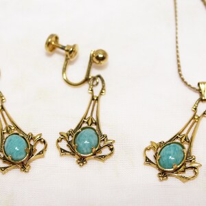 May include: A vintage jewelry set featuring a necklace and clip-on earrings. The set has a gold-tone metal finish with a decorative filigree design. Each piece holds a light blue oval stone. The earrings are clip-on style.