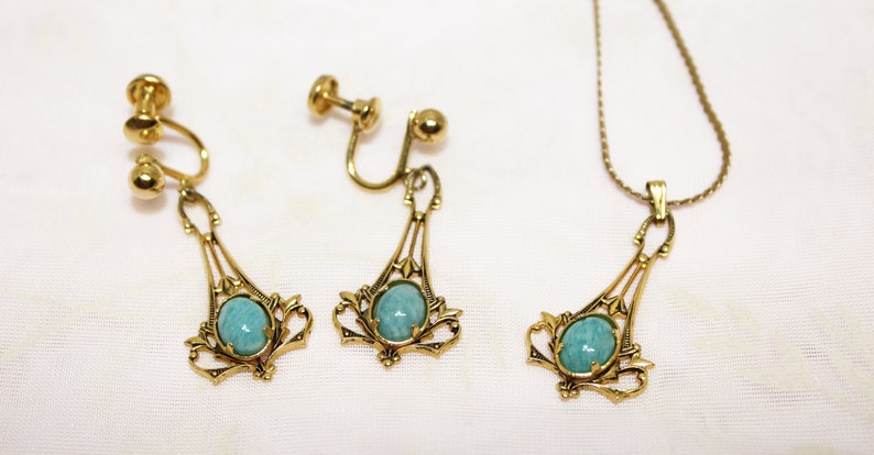 May include: A vintage jewelry set featuring a necklace and clip-on earrings. The set has a gold-tone metal finish with a decorative filigree design. Each piece holds a light blue oval stone. The earrings are clip-on style.