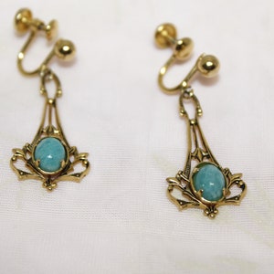 May include: A pair of vintage clip-on earrings. Each earring features a gold-toned filigree design with an oval, light blue gemstone. The earrings have a screw-back mechanism for secure wear. The jewelry is displayed on a white fabric.