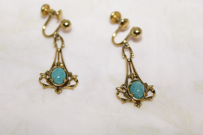 May include: A pair of vintage clip-on earrings. Each earring features a gold-toned filigree design with an oval, light blue gemstone. The earrings have a screw-back mechanism for secure wear. The jewelry is displayed on a white fabric.