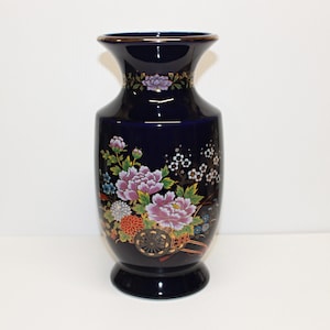 May include: A dark blue ceramic vase with a gold rim and a flared top. The vase features a floral design with pink peonies, a small cart, and other colourful flowers. The vase is approximately 25 cm tall.