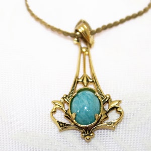 May include: A gold-toned pendant necklace featuring an oval, light blue gemstone. The pendant has an ornate, Art Nouveau-inspired design with delicate filigree details. The necklace chain is a thin, twisted rope style.