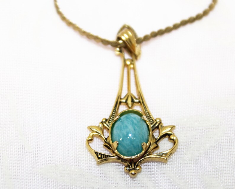 May include: A gold-toned pendant necklace featuring an oval, light blue gemstone. The pendant has an ornate, Art Nouveau-inspired design with delicate filigree details. The necklace chain is a thin, twisted rope style.