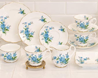 May include: A white porcelain tea set featuring a delicate blue forget-me-not floral pattern and gold-trimmed scalloped edges. The collection includes plates, saucers, teacups, a creamer pitcher, a sugar bowl, and a small square dish.