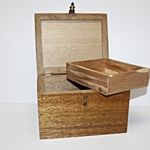 May include: A wooden chest with a hinged lid and a small, removable tray. The chest is a warm, golden brown colour with a textured surface. The lid is open, revealing the interior.