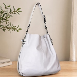 May include: A white leather shoulder bag with a single strap and silver-tone hardware. The bag has a slouchy design and black trim. The bag is sitting on a wooden surface.