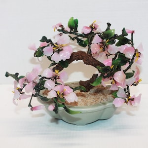 May include: A decorative bonsai tree sculpture featuring a textured brown trunk, dark green leaves, and light pink flower petals with yellow centers. The tree is set in a light gray, scalloped-edge ceramic pot filled with small tan pebbles.