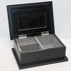 May include: A black wooden jewelry box with a hinged lid, open to reveal a mirrored interior and gray felt-lined compartments. The box is rectangular with a raised base and silver hinges.