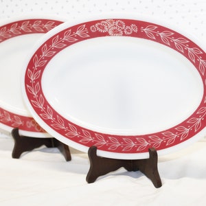 May include: Two white oval serving platters with a red border featuring a leaf and floral design. The platters are displayed on dark brown wooden stands, showcasing a vintage aesthetic.