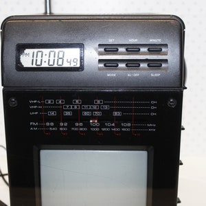 May include: A black and silver portable television with a built-in clock and radio. The clock displays "10:08 49 PM". The radio has a dial with VHF-L, VHF-H, UHF, FM, and AM bands.