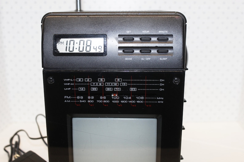 May include: A black and silver portable television with a built-in clock and radio. The clock displays "10:08 49 PM". The radio has a dial with VHF-L, VHF-H, UHF, FM, and AM bands.