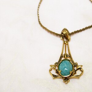 May include: A gold-toned pendant necklace with a round, light blue gemstone. The pendant has an ornate, Art Deco-inspired design. A second, simpler gold chain is also visible.