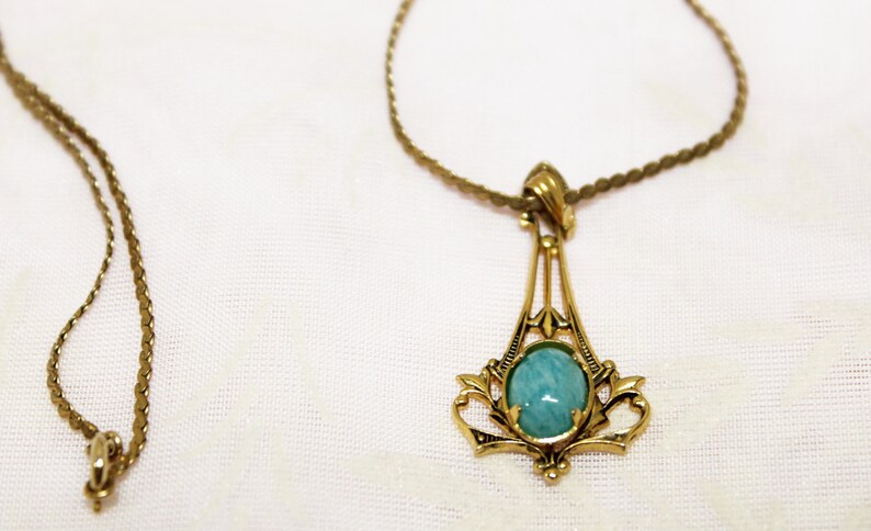 May include: A gold-toned pendant necklace with a round, light blue gemstone. The pendant has an ornate, Art Deco-inspired design. A second, simpler gold chain is also visible.