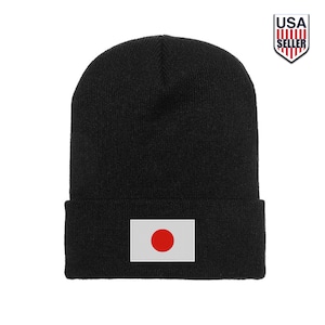 May include: A solid black knit beanie featuring a rectangular patch on the front cuff. The patch displays the national flag of Japan, which consists of a red circle centered on a white rectangular background. The beanie has a ribbed texture.
