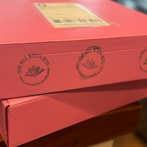 Pode incluir: Duas caixas cor-de-rosa empilhadas uma em cima da outra. A caixa de cima tem um rótulo castanho e um carimbo preto que diz "The Full Book Box" com um desenho floral. A caixa de baixo tem o mesmo carimbo no lado.