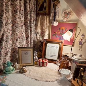 May include: A collection of vintage decor items including a floral curtain, a framed sign reading "Sing a song of vintage", a brass lamp with a floral shade, a white lace doily, a green glass pitcher, a red and white striped tin, and amber glass.