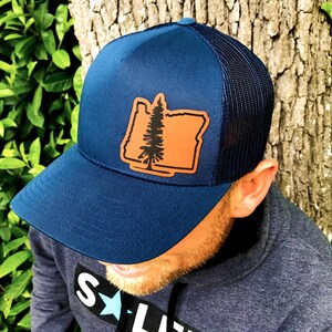 May include: A navy blue trucker hat with a brown leather patch featuring the outline of the state of Oregon and a pine tree. The hat has a black mesh back.