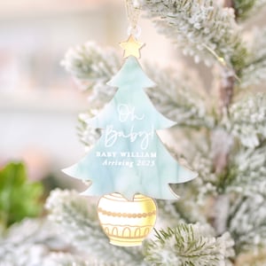 Op de afbeelding: Een lichtblauwe kerstboomversiering met een gouden ster bovenop. De versiering heeft de tekst "Oh Baby!" en "Baby William Arriving 2023" erop. De versiering hangt aan een besneeuwde kerstboom.