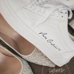 Puede incluir: Zapatillas de lona blancas con cordones, con la inscripción "Mrs. Castillo" en escritura negra. La imagen también muestra un par de tacones de punta adornados. Las zapatillas son de estilo clásico, adecuadas para uso casual.