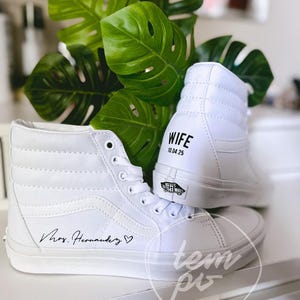 K&ouml;nnte beinhalten: Ein Paar wei&szlig;e High-Top-Sneaker mit dem Schriftzug "Mrs. Hernandez" und einem Herzsymbol an der Seite. Die Ferse eines Sneakers zeigt den Text "WIFE" &uuml;ber dem Datum "10.04.25" und ein schwarzes "Vans Off The Wall"-Logo an der Sohle.