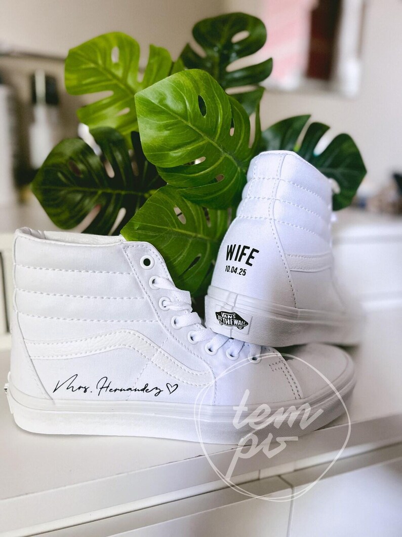 K&ouml;nnte beinhalten: Ein Paar wei&szlig;e High-Top-Sneaker mit dem Schriftzug "Mrs. Hernandez" und einem Herzsymbol an der Seite. Die Ferse eines Sneakers zeigt den Text "WIFE" &uuml;ber dem Datum "10.04.25" und ein schwarzes "Vans Off The Wall"-Logo an der Sohle.
