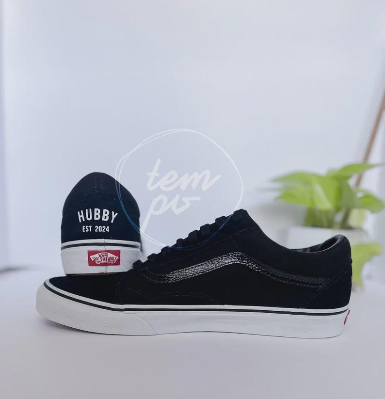May include: A pair of black low-top trainers with white rubber soles and laces. The heel of one shoe features the white text HUBBY EST 2024 above a red Vans Off The Wall logo. The side of the shoe displays a black textured side stripe.