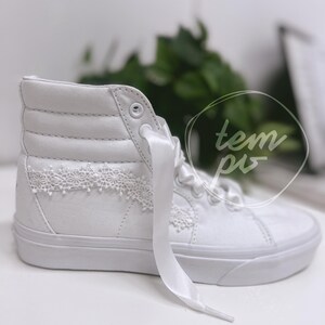 K&ouml;nnte beinhalten: Ein wei&szlig;er High-Top-Sneaker mit glattem Obermaterial aus Canvas, einer dicken Gummisohle und breiten Satinband-Schn&uuml;rsenkeln. Das Seitenteil ist mit einem zarten, wei&szlig;en Spitzenbesatz in Blumenoptik verziert. Der Text "tem pis" ist zu sehen.