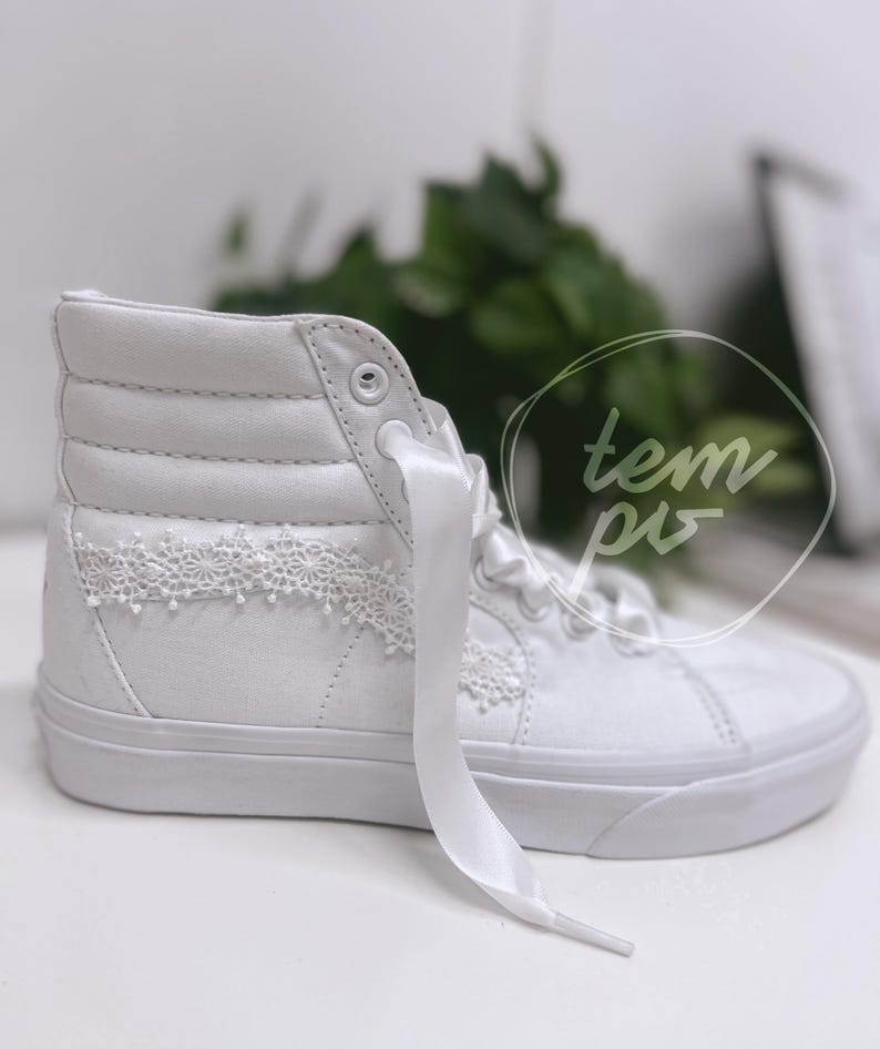 K&ouml;nnte beinhalten: Ein wei&szlig;er High-Top-Sneaker mit glattem Obermaterial aus Canvas, einer dicken Gummisohle und breiten Satinband-Schn&uuml;rsenkeln. Das Seitenteil ist mit einem zarten, wei&szlig;en Spitzenbesatz in Blumenoptik verziert. Der Text "tem pis" ist zu sehen.
