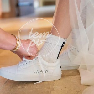 K&ouml;nnte beinhalten: Ein wei&szlig;er Sneaker mit wei&szlig;en Schn&uuml;rsenkeln und schwarzem Schriftzug Mrs. Hury, gefolgt von einem kleinen Herzsymbol. Der Sneaker wird mit einer wei&szlig;en, gerippten Socke getragen, die zwei d&uuml;nne schwarze Streifen am oberen B&uuml;ndchen aufweist.