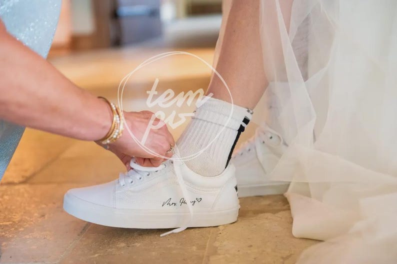 K&ouml;nnte beinhalten: Ein wei&szlig;er Sneaker mit wei&szlig;en Schn&uuml;rsenkeln und schwarzem Schriftzug Mrs. Hury, gefolgt von einem kleinen Herzsymbol. Der Sneaker wird mit einer wei&szlig;en, gerippten Socke getragen, die zwei d&uuml;nne schwarze Streifen am oberen B&uuml;ndchen aufweist.