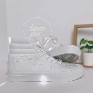K&ouml;nnte beinhalten: Ein Paar kn&ouml;chelhohe Sneaker in reinem Wei&szlig; mit einem strukturierten, glitzernden Seitenstreifen und wei&szlig;en Satinband-Schn&uuml;rsenkeln. Die Schuhe haben eine dicke Gummisohle und eine hohe Silhouette, mit dem Wort "tempo" in einem kreisf&ouml;rmigen Logo.