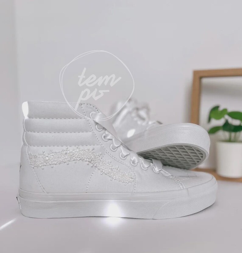 K&ouml;nnte beinhalten: Ein Paar kn&ouml;chelhohe Sneaker in reinem Wei&szlig; mit einem strukturierten, glitzernden Seitenstreifen und wei&szlig;en Satinband-Schn&uuml;rsenkeln. Die Schuhe haben eine dicke Gummisohle und eine hohe Silhouette, mit dem Wort "tempo" in einem kreisf&ouml;rmigen Logo.