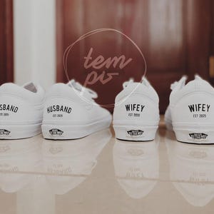 May include: Four white canvas sneakers, two labeled "HUSBAND EST 2020" and two labeled "WIFEY EST 2020". The shoes are arranged in pairs, with the backs of the shoes facing the camera. The shoes are on a reflective surface.