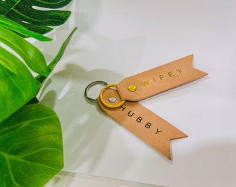 May include: A leather keychain with two tags. One tag says "WIFEY" in gold lettering and the other tag says "HUBBY" in black lettering.