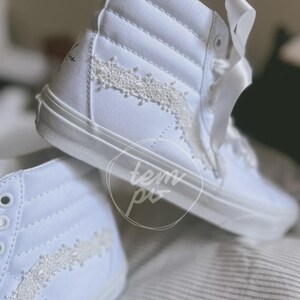 K&ouml;nnte beinhalten: Ein Paar wei&szlig;e High-Top-Sneaker mit einem Obermaterial aus Segeltuch, wei&szlig;en Satinband-Schn&uuml;rsenkeln und einer zarten wei&szlig;en Spitzenborte entlang des Seitenteils. Die Sneaker haben eine hohe Silhouette mit verst&auml;rkter Ferse und einer durchgehenden wei&szlig;en Gummisohle.