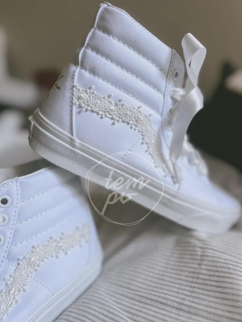 K&ouml;nnte beinhalten: Ein Paar wei&szlig;e High-Top-Sneaker mit einem Obermaterial aus Segeltuch, wei&szlig;en Satinband-Schn&uuml;rsenkeln und einer zarten wei&szlig;en Spitzenborte entlang des Seitenteils. Die Sneaker haben eine hohe Silhouette mit verst&auml;rkter Ferse und einer durchgehenden wei&szlig;en Gummisohle.