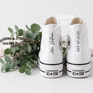 May include: A pair of white high-top canvas sneakers with black trim and rubber soles. The left shoe features the text Mrs. Sullivan, and the right shoe features the text EST 07.23.25 printed vertically on the heel. The Converse All Star logo is visible.