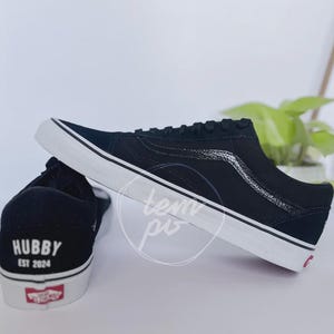 May include: A pair of black low-top trainers with white rubber soles. The heel of one shoe features the white text "HUBBY" above "EST 2024". The side of the shoe displays a black textured side stripe, and a red logo tag is on the heel sole.