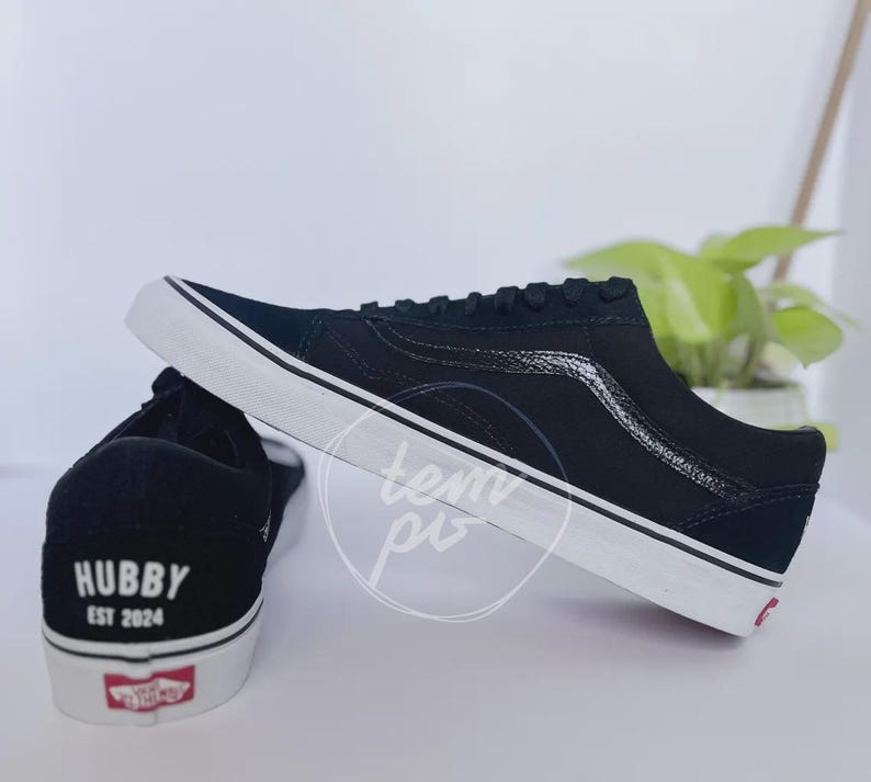 May include: A pair of black low-top trainers with white rubber soles. The heel of one shoe features the white text "HUBBY" above "EST 2024". The side of the shoe displays a black textured side stripe, and a red logo tag is on the heel sole.