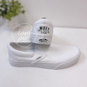 May include: White Vans slip-on sneakers. The heel of one shoe is visible, with the word "Wifey", the name "Mrs. Beauty", and the date "10.19.2024" printed in black. The Vans logo is also visible.