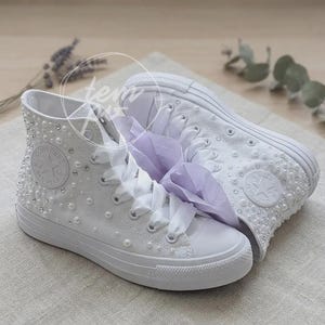May include: A pair of white high-top sneakers featuring a canvas upper adorned with scattered white pearls and clear rhinestones. The shoes include white rubber toe caps, matching white rubber soles, and wide white ribbon laces with a light purple fabric accent.